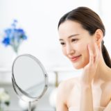 A woman puts her hand on her cheek and looks at her skin condition in the mirror