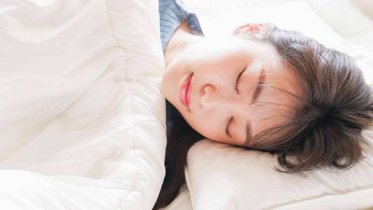 A young woman sleeping in bed. She is wrapped in a white blanket and resting her head on a white pillow with a calm expression and closed eyes.