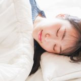 A young woman sleeping in bed. She is wrapped in a white blanket and resting her head on a white pillow with a calm expression and closed eyes.