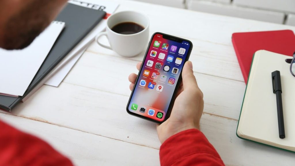 机の上にあるノートやコーヒーカップの近くでスマートフォンを持って操作している様子。スマートフォンの画面にはアプリのアイコンが並んでいる。