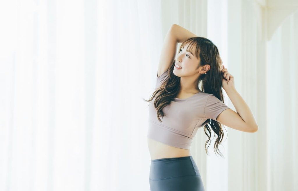 若い女性がストレッチをしている様子。フィットネスウェアを着て、明るい背景の中でリラックスした表情で腕を上げている。
