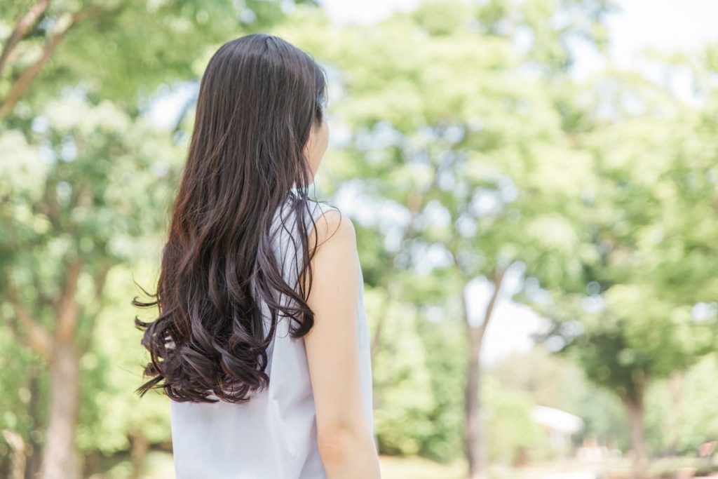 長いウェーブヘアの女性が屋外で、背中をカメラに向けて立ち、緑の木々に囲まれている。