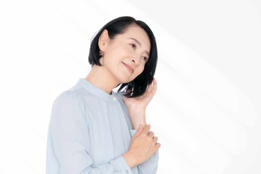 短い黒髪の中年女性が、髪に触れながら優しく微笑んでいる。ライトブルーのブラウスを着ている。
