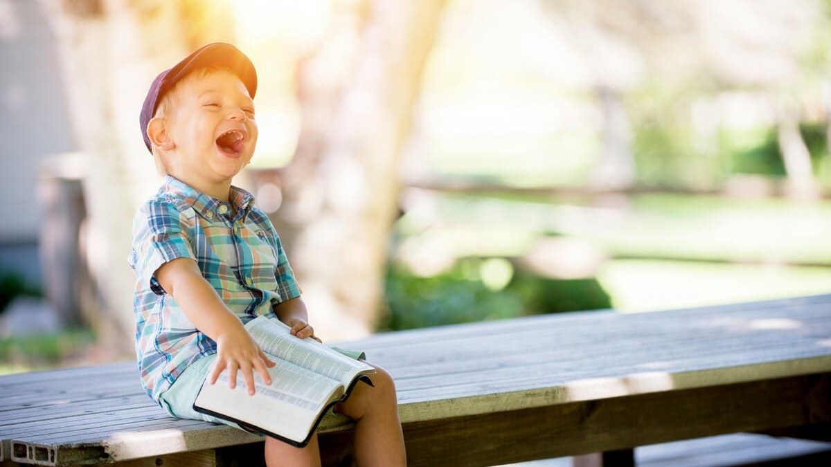 A young boy wearing a cap and a plaid shirt sits on a wooden table, laughing loudly. He is holding a Bible in his hands.