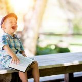 A young boy wearing a cap and a plaid shirt sits on a wooden table, laughing loudly. He is holding a Bible in his hands.