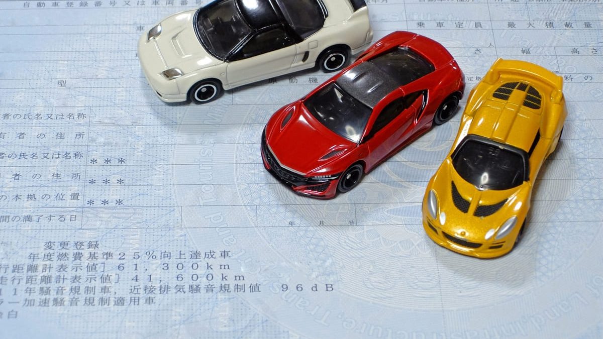 Three toy cars—one white, one red, and one yellow—placed on top of a Japanese vehicle inspection document.