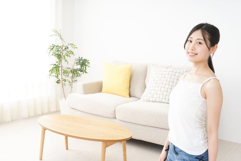 明るいリビングルームで笑顔の若い女性。背景にはベージュのソファ、装飾用のクッション、木製のコーヒーテーブル、観葉植物がある。