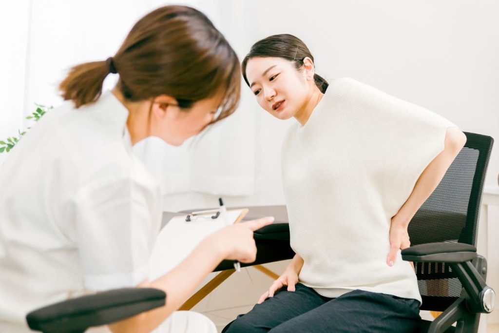 黒いオフィスチェアに座り、腰に手を当てて痛みを訴える女性が、白衣の医療従事者と相談している様子。医療従事者はクリップボードを指さしています。