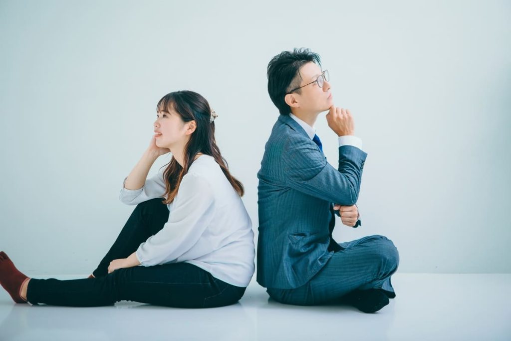 スーツを着た男性とカジュアルな服装の女性が背中合わせに座り、考え事をしているように見える。背景は淡い青色。