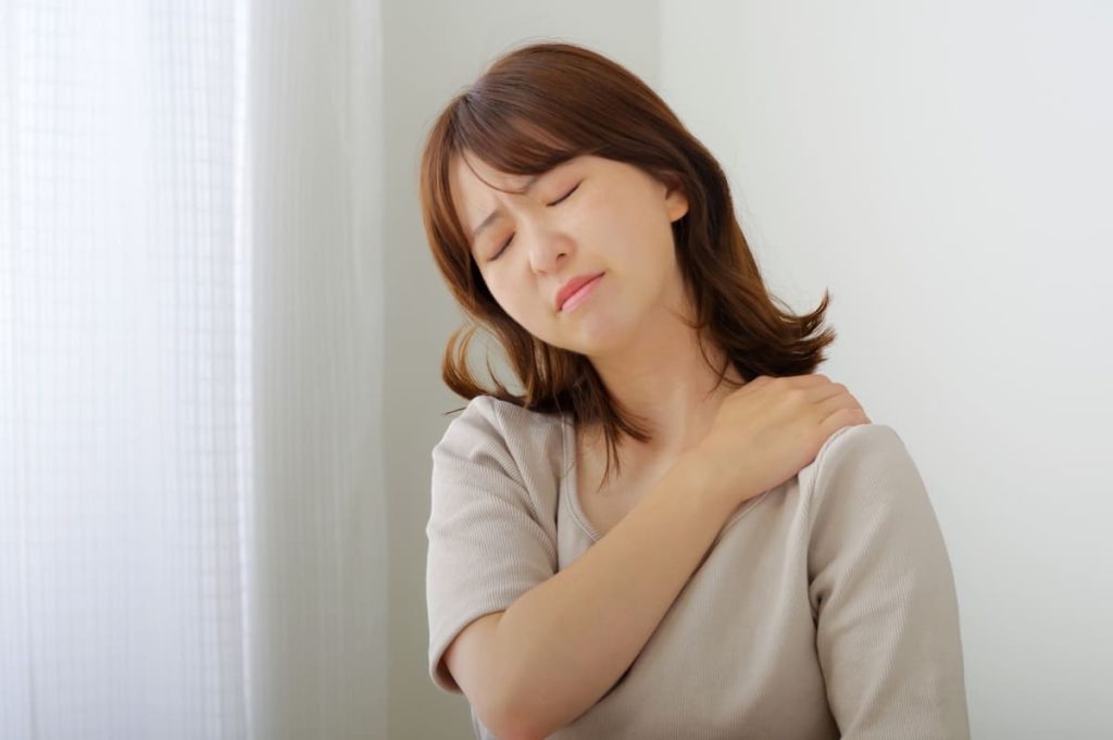 窓際の明るい部屋で肩を押さえ、不快そうな表情をしている女性。