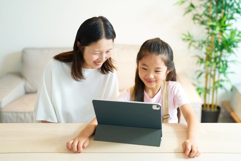 母親と娘が明るいリビングルームで一緒にテーブルに座り、タブレットを見ながら微笑んでいる。