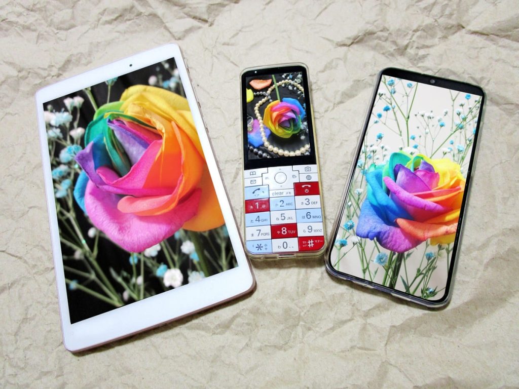 テクスチャのある背景に、タブレット、ガラケー、スマートフォンの3つのデバイスが並べられ、それぞれの画面にカラフルな虹色のバラが表示されています。