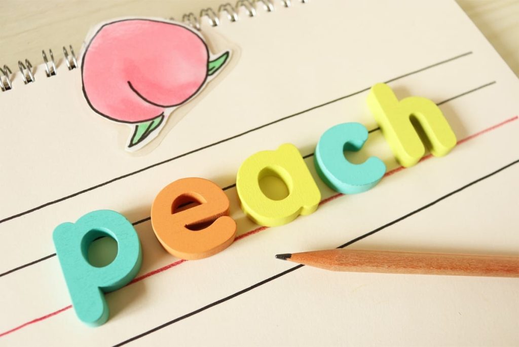 ノートにカラフルなスポンジ文字で「peach」と綴られており、隣には桃の絵と木製の鉛筆が置かれている。