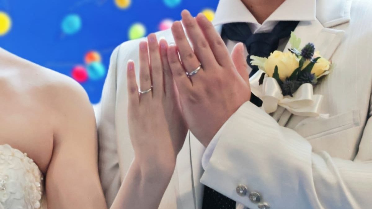 A close-up of a bride and groom showing their wedding rings by raising their hands side by side. The groom is wearing a white suit with a yellow and purple boutonniere, while the bride is in a strapless dress. The background features colorful balloons.