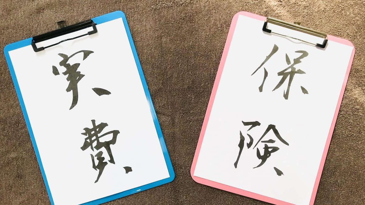 Two clipboards placed on a brown surface, each displaying Japanese calligraphy. The clipboard with a blue border reads "実費" (actual expenses), and the one with a pink border reads "保険" (insurance).
