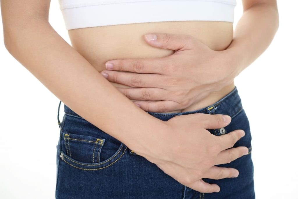 白いトップスと青いジーンズを着た人が両手でお腹を押さえている様子。不快感や痛みを示唆している。