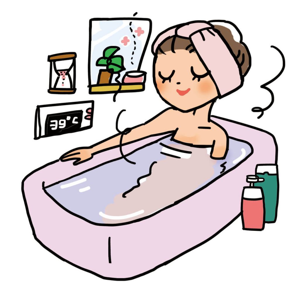 ピンク色のタオルを頭に巻いた女性が、39°Cに設定された温かいお風呂に浸かってリラックスしています。背景には鏡、小さな観葉植物、入浴用品が見えます。