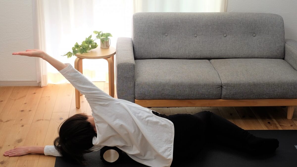 A person lying on their side on a black yoga mat, using a foam roller for stretching in a bright living room with a gray sofa and a small wooden table with plants.