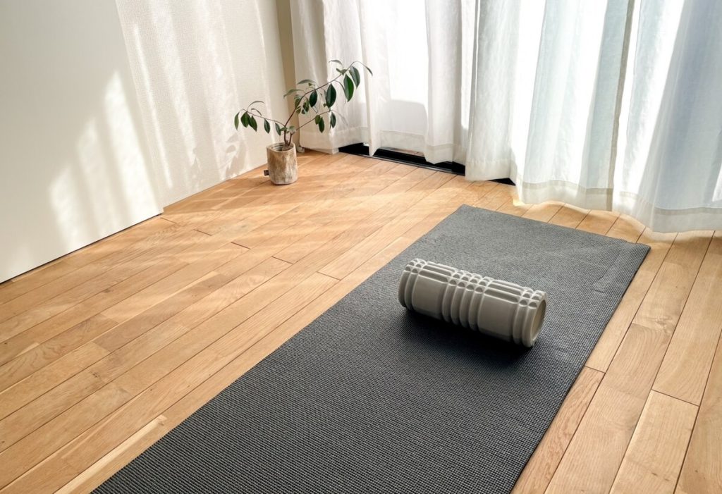明るい部屋で、ヨガマットの上に置かれた灰色のフォームローラー。木製の床と白いカーテン付きの窓、近くには鉢植えの植物がある。