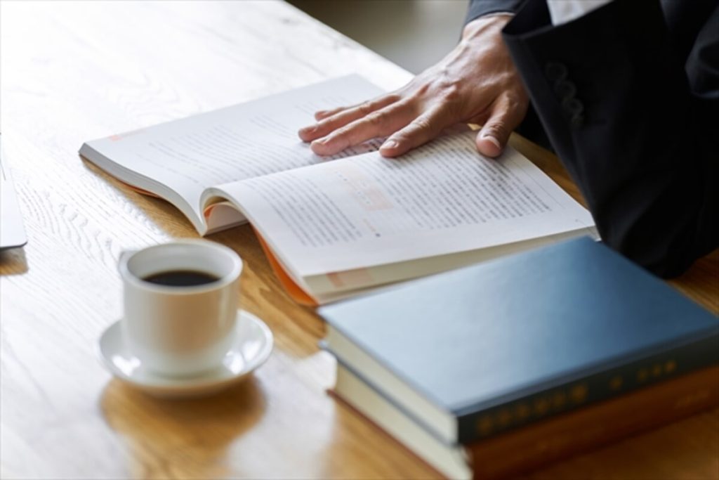スーツを着た人物が木製のテーブルで開いた本を読んでいる様子。近くにはコーヒーカップと閉じた本が2冊置かれている。