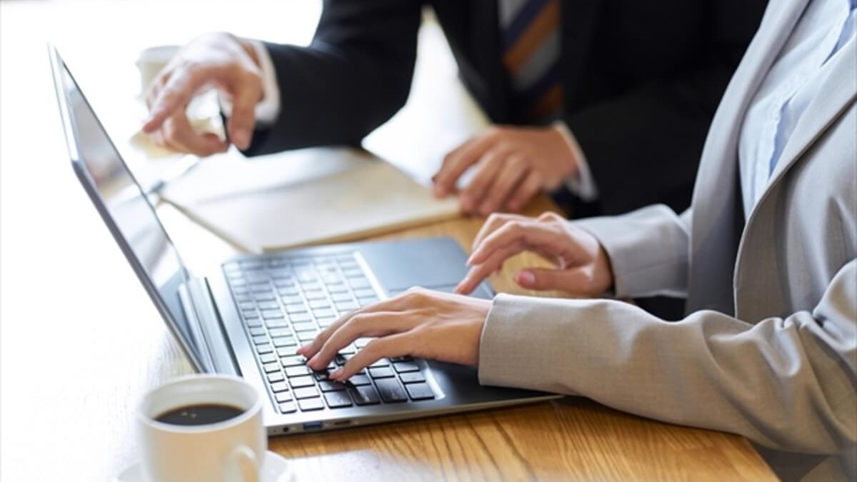 Two people in business attire working at a wooden table. One person is typing on a laptop, while the other is pointing at the screen. A cup of coffee is on the table.