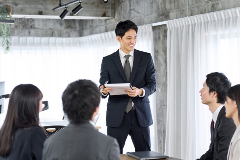 スーツを着た男性が立ちながら書類を持ち、微笑んで座っている同僚たちに話しかけている。モダンな会議室内の様子。