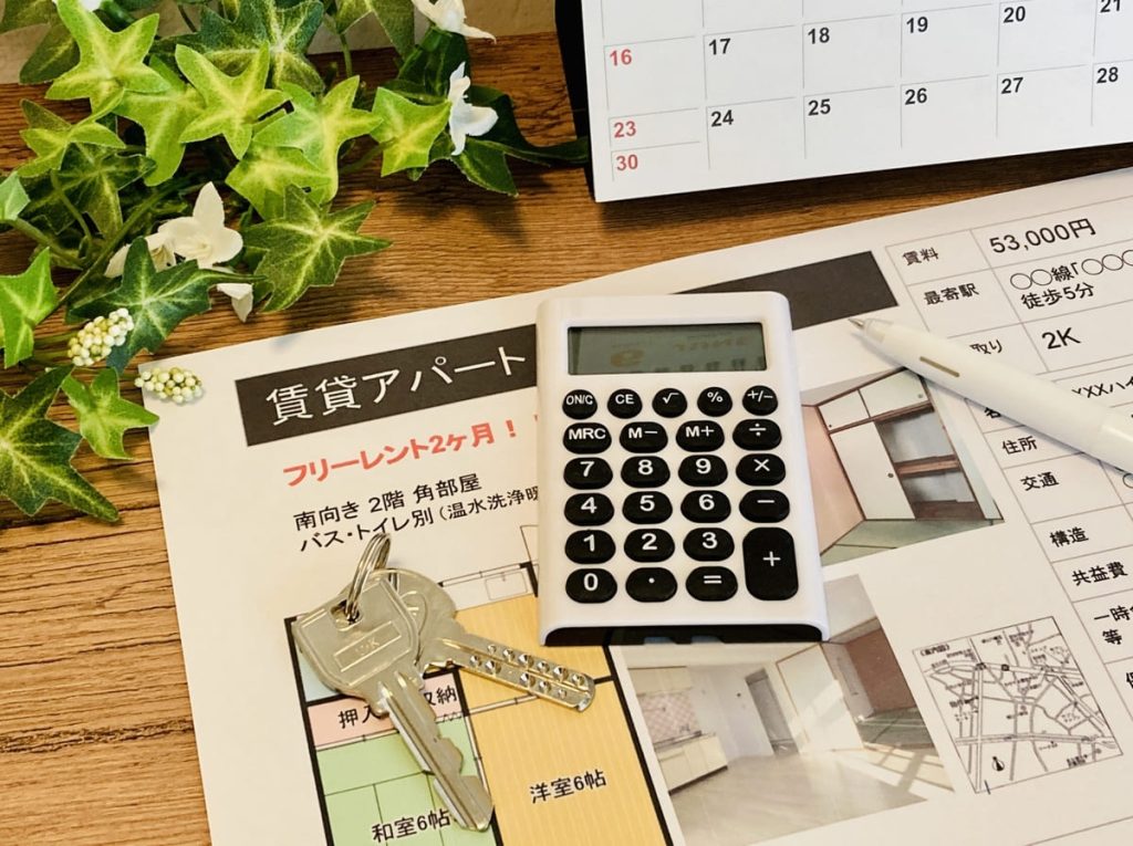 電卓、鍵、ペンが置かれた賃貸物件のチラシ。背景には小さな観葉植物とカレンダーが見えます。チラシには「フリーレント2ヶ月」の賃貸アパート情報が記載されています。
