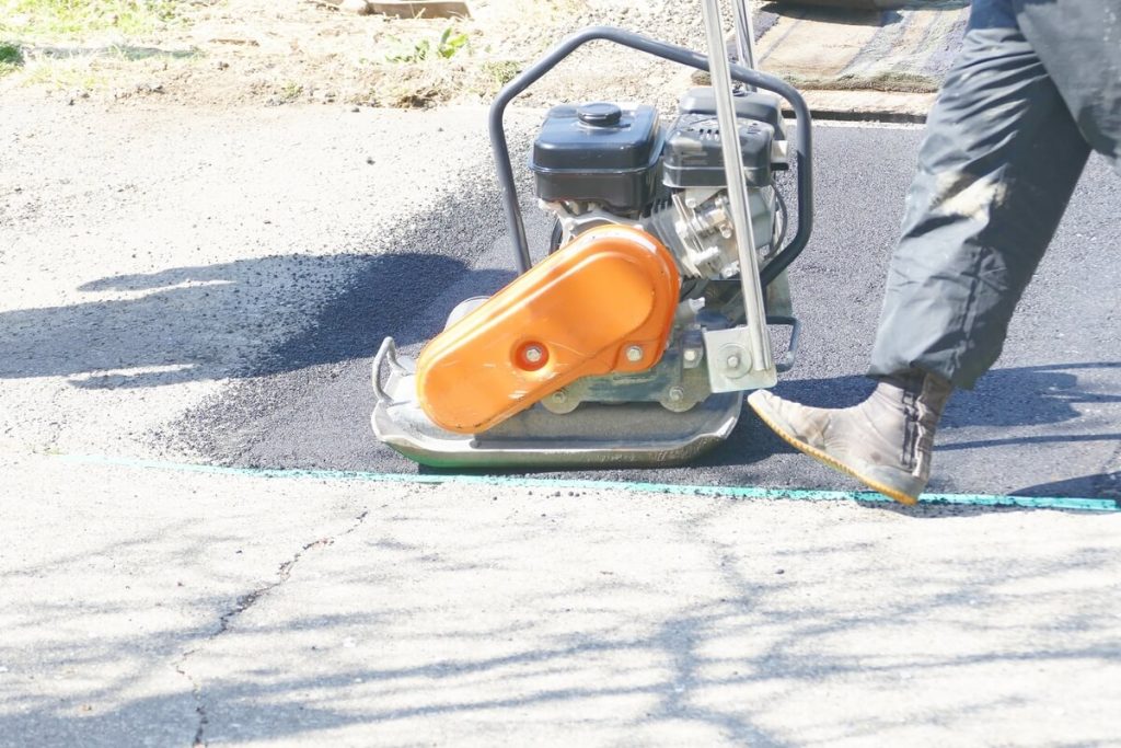 新しく舗装されたアスファルトの上でプレートコンパクターを操作する作業員。オレンジ色のエンジンカバーと周囲の影が見えます。