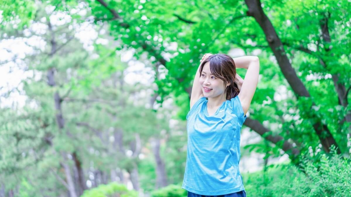 A cheerful woman in a light blue sports outfit stretching her arms over her head in a lush green outdoor setting with trees in the background.