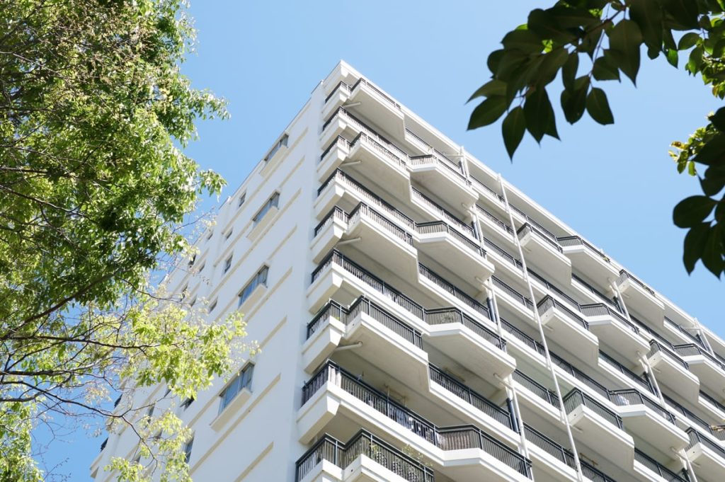 晴れた日に下から見上げた白い現代的なアパートの建物。複数のバルコニーがあり、左右に緑の木の枝が写っています。