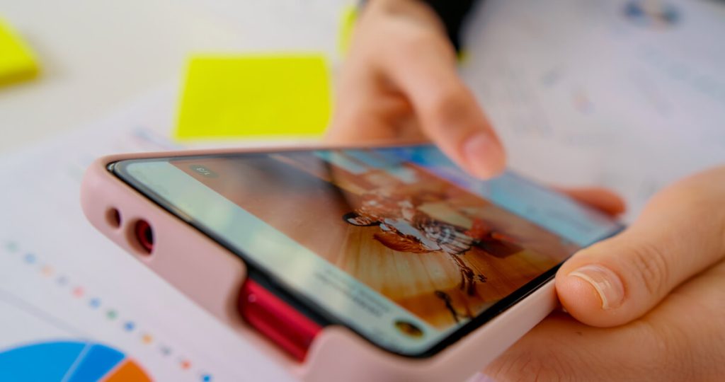 ピンク色のケースに入ったスマートフォンを手に持つクローズアップ画像。スマートフォンには画像または動画が表示されており、背景にはグラフ付きの書類と黄色の付箋が見える。