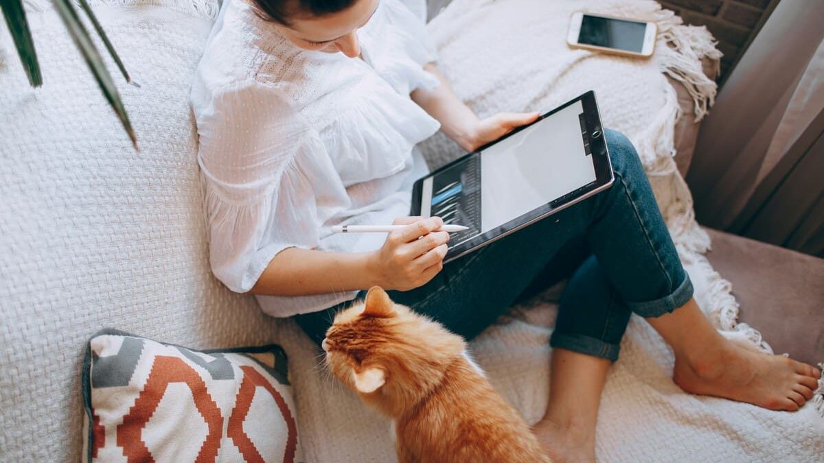 A woman sitting on a sofa, using a digital pen on a tablet, with an orange cat nearby looking at her. A smartphone rests on the sofa beside her.