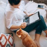 A woman sitting on a sofa, using a digital pen on a tablet, with an orange cat nearby looking at her. A smartphone rests on the sofa beside her.