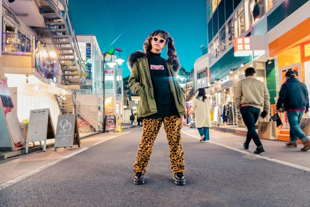 緑のパーカー、ヒョウ柄のパンツ、サングラスを身に着けたスタイリッシュな人物が、カラフルな都市の夜の街中で自信を持って立っている。街には店舗が並び、鮮やかな照明が灯っている。