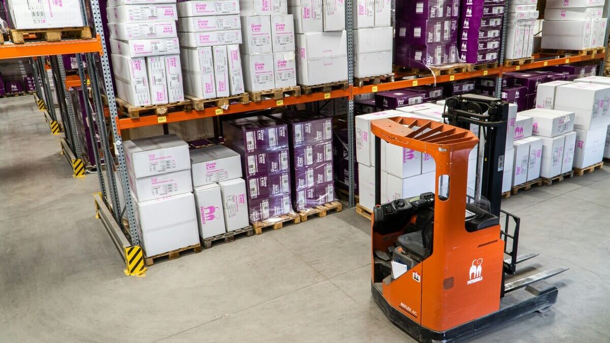 A warehouse interior with shelves stacked with boxes and an orange forklift parked in the foreground. The shelves are filled with white and purple-labeled packages.