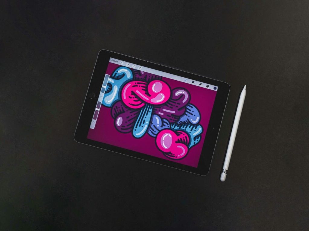 カラフルなデジタルイラストが表示されたタブレットと、その横に置かれたスタイラスペン。背景は暗い色です。