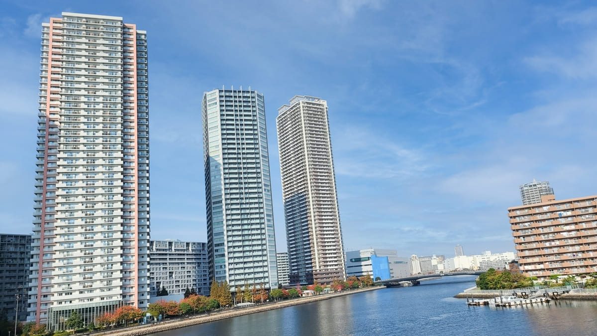 A view of three tall modern apartment buildings by a river, with clear blue skies in the background. Trees and smaller buildings line the riverbank.