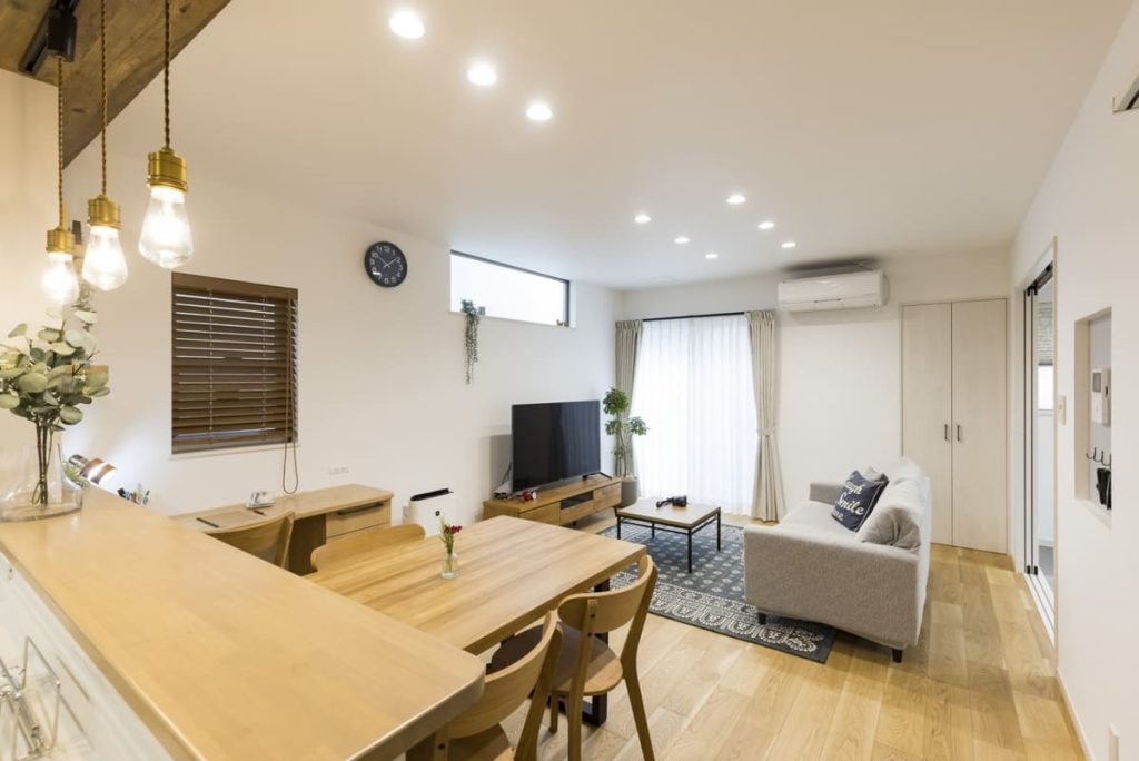 木製の床、ダイニングテーブルと椅子、ソファ、テレビがあるモダンなリビングルーム。カーテンから自然光が差し込む明るい室内で、ダイニングエリアにはペンダントライトが吊り下げられています。