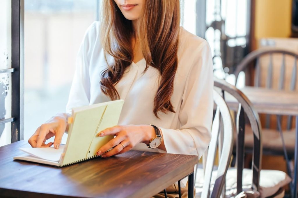 カフェで木製のテーブルに座り、ノートに目を通すまたは書き込む白いブラウスの女性。窓から差し込む陽光が明るく穏やかな雰囲気を演出しています。