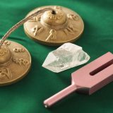 A set of Tibetan tingsha cymbals, a clear quartz crystal, and a pink tuning fork arranged on a green fabric background.