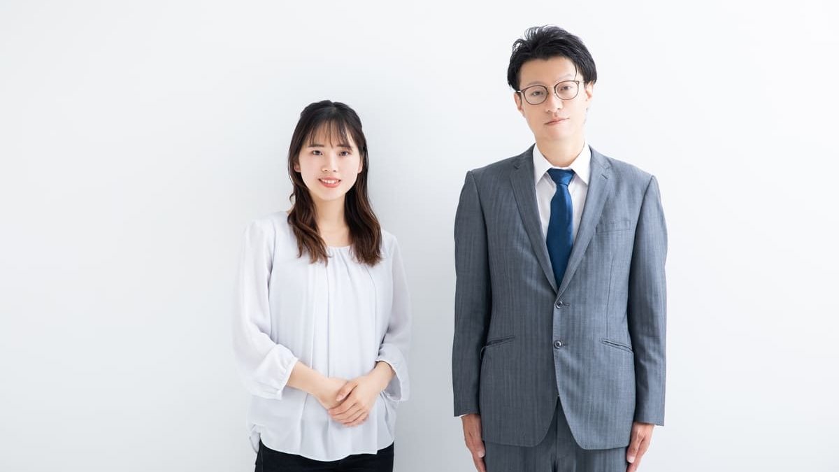 A man in a gray suit and a woman in a white blouse standing in front of a plain white background. Both are smiling lightly and facing forward.