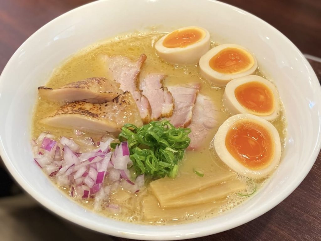 炙り鶏、半熟卵、刻み赤玉ねぎ、メンマ、ネギが乗った濃厚な鶏白湯ラーメンが、白い器に盛られています。