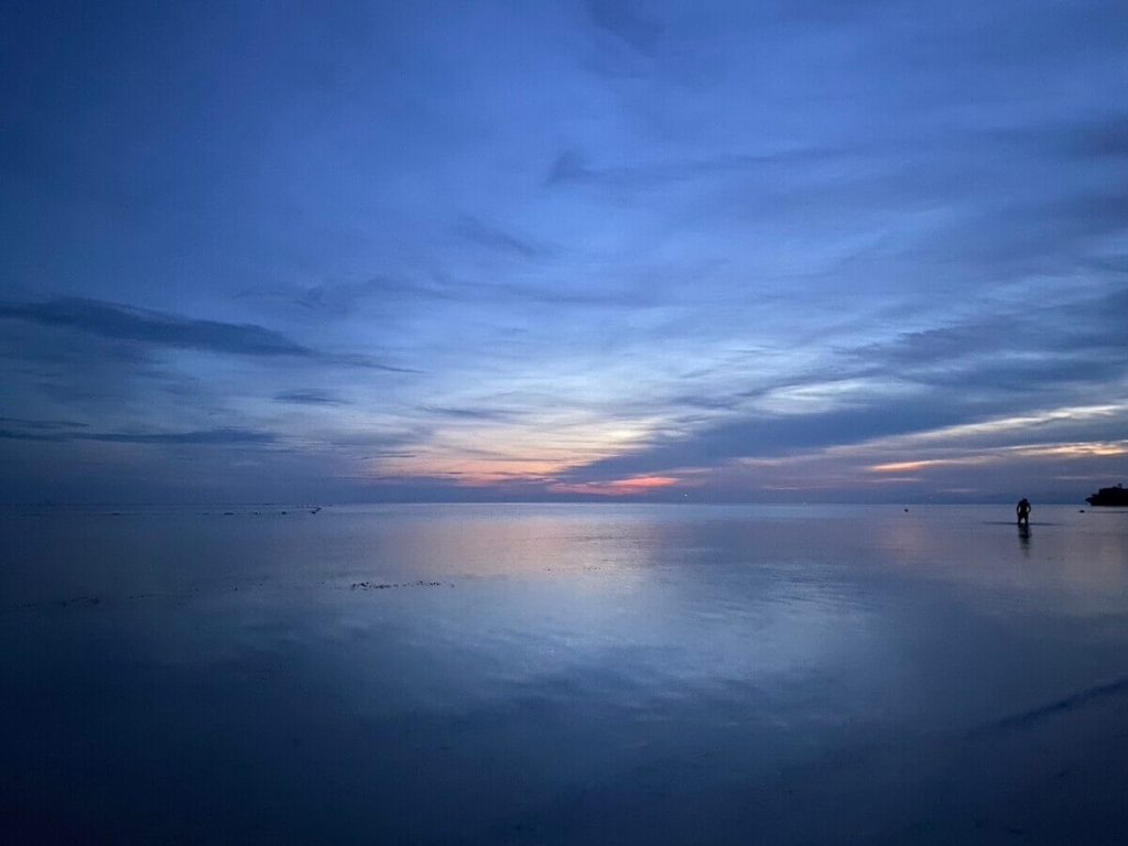 夕暮れの穏やかな海の風景。青とオレンジの淡い空の色が静かな水面に映り込んでいます。遠くの海岸近くに人影が見えます。