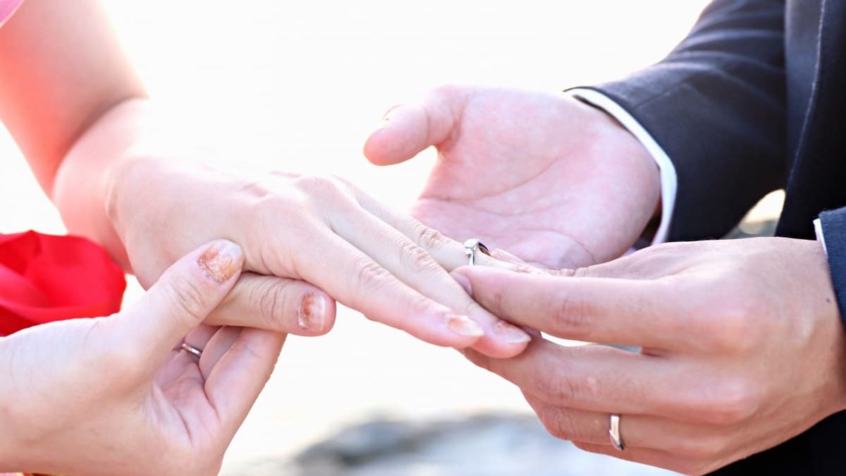A close-up of a couple exchanging rings during a wedding ceremony. The bride's hand is being gently held as the groom places a ring on her finger.