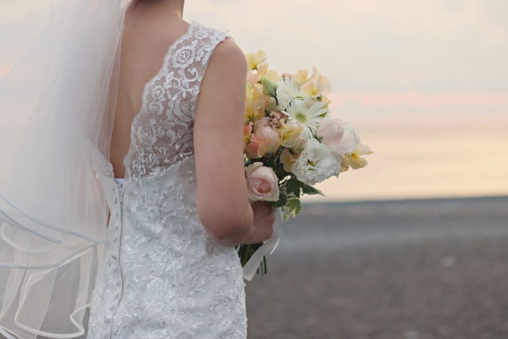 レースのウェディングドレスを着た花嫁がパステルカラーの花束を持ち、海と柔らかな夕日を背景に立っています。