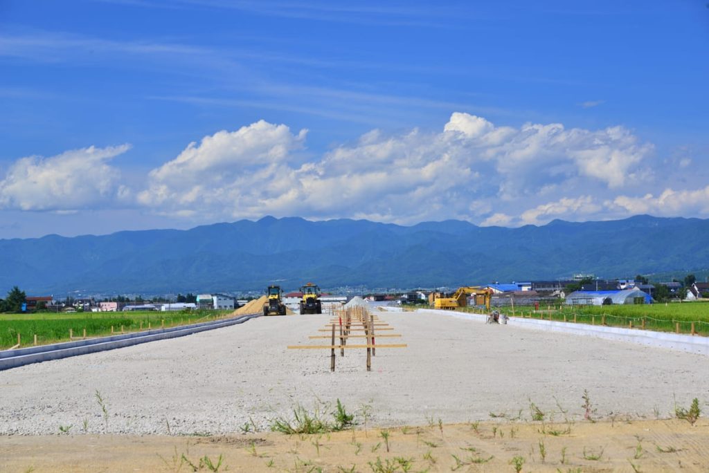 青空の下、山々を背景に進行中の道路工事現場。中央には未舗装の道路が伸び、工事用の木枠が並んでいる。重機や作業車が配置され、周囲には田畑や建物が点在している。