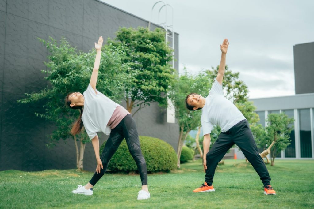 屋外の芝生の上でストレッチをする中年の男女。二人とも白いTシャツと黒いパンツを着用し、片手を上げて体を横に倒すポーズをとっている。緑の木々とモダンな建物が背景にあり、リラックスした雰囲気が感じられる。
