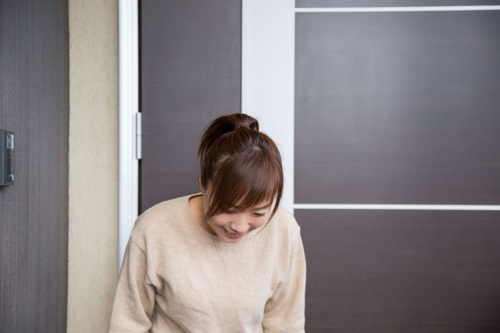 玄関先で笑顔でお辞儀をする女性。ベージュのセーターを着ており、礼儀正しく挨拶している様子。背景にはモダンなデザインの玄関ドアが見える。