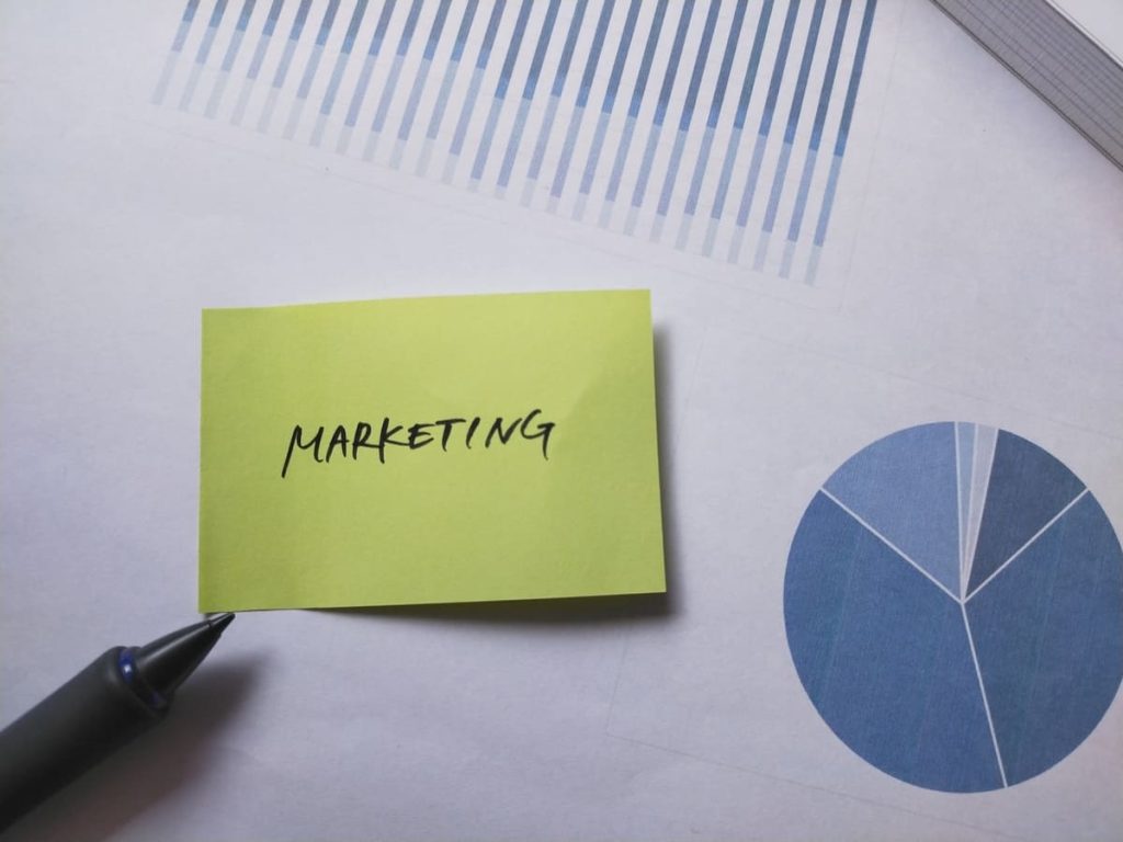 「MARKETING」と書かれた黄緑色の付箋が、グラフやデータが印刷された紙の上に置かれている。左下には黒いペンが見える。