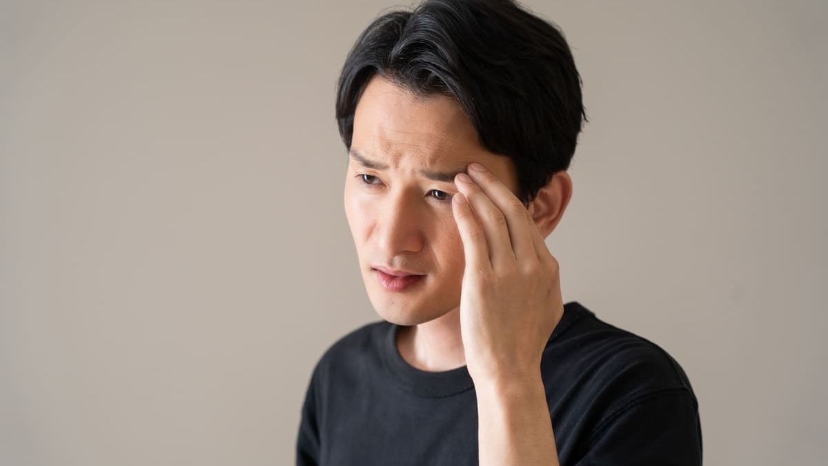 A man in a black T-shirt touches his temple with a worried expression, appearing to have a headache or be in deep thought. The background is a plain beige wall.